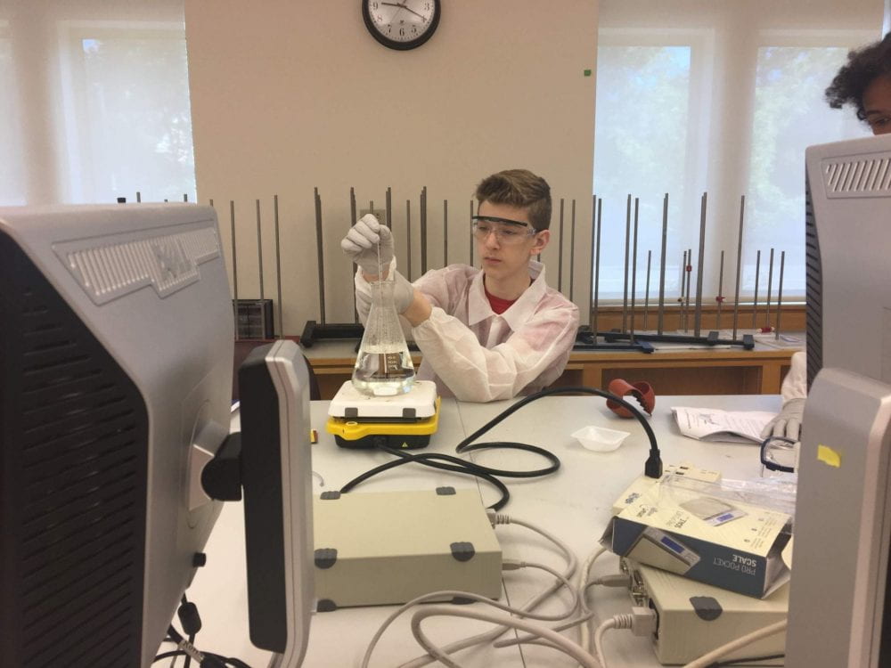 A scholar wearing goggles and a white lab coat is seated at a table. They are working with a variety of equipment in a laboratory setting. 