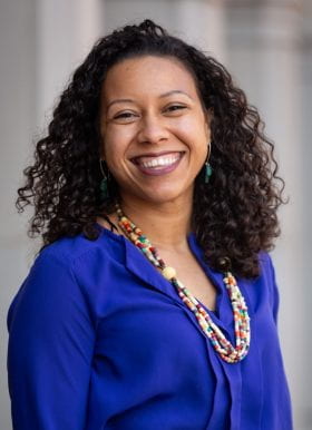 Photo of Ashley Prince, Program Coordinator, standing in front of a blurred gray background. Ashley is smiling, has medium length curly dark brown hear, and is wearing a royal blue blouse and colorful beaded necklace.