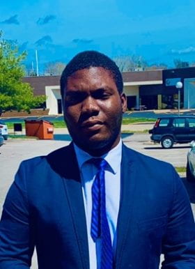 Photo of Philip Lawal, Program Assistant. Philip is standing outside with a building and parking lot behind him. He has short cropped black hair and is wearing a dark blue blazer over a light blue button-down shirt with a dark blue necktie featuring a Windsor knot.