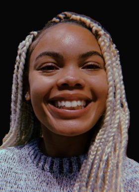 Photo of Sparkle Whittaker, Program Assistant. Sparkle is in front of a black background and has platinum blonde braids. Sparkle is smiling and wearing a gray sweater.