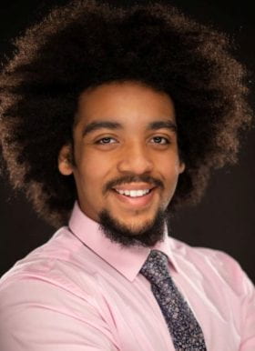 Photo of Carlos Tyse, Program Assistant. Carlos is smiling in front of a black background, and has facial hair and natural hair. Carlos wears a pink button-down shirt and a gray and pink patterned necktie featuring a Windsor knot