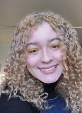 Photo of Jayla Coleman, Program Assistant. Jayla is indoors, smiling, and has burly blonde hair. Jayla is wearing a black turtleneck and denim overalls.