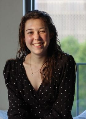 Photo of Natalie Fisher, Program Assistant. Natalie is seated indoors in front of a window, is smiling, has long brown hair worn loose, and is wearing a black three quarter sleeve length top with small white polka-dots.