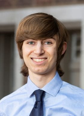 Photo of Matthew Wagner, Program Assistant. Matthew is standing outside of a brick building. Matthew is in focus, and the background is blurred. Matthew has brown hair, is smiling, and wears a light blue button-down shirt with a dark blue necktie featuring a Windsor knot.
