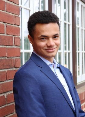 Photo of Christian Alexander, Program Assistant, standing outdoors in front of a brick building with a white framed window. He is smiling and wears a dark blue blazer over a light blue button-down shirt.