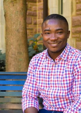 Photo of Peter Ogunniran, Graduate Fellow, smiling while seated outdoors. He has close cropped hair, neatly trimmed facial hair, and is wearing a red, white, and blue checked shirt. He sits on a bench beneath a tree in front of a stone building.