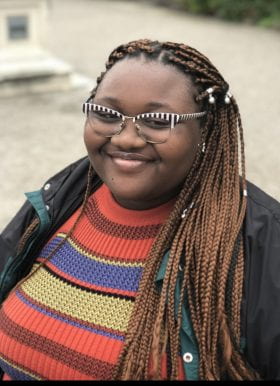 Photo of Porscha Hayes, Program Assistant, standing outdoors. She is smiling, has her hair in long braids, is wearing black and white vertical striped glasses and a horizonal striped shirt in red tones, yellow, blue, and black, beneath an unbuttoned black jacket with hunter green trim.
