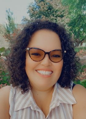 Photo of Jennifer Ramirez, Administrative Coordinator. Jennifer is outdoors in front of a tree and has dark curly, shoulder-length hair worn down, tortoise shell glasses, and a sleeveless collared top with white and tan stripes.