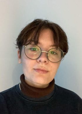 Program Assistant Jacquelyn Grimes is pictured in front of a gray background. They have short dark hair and are wearing round metal frame glasses and a dark turtle-neck.